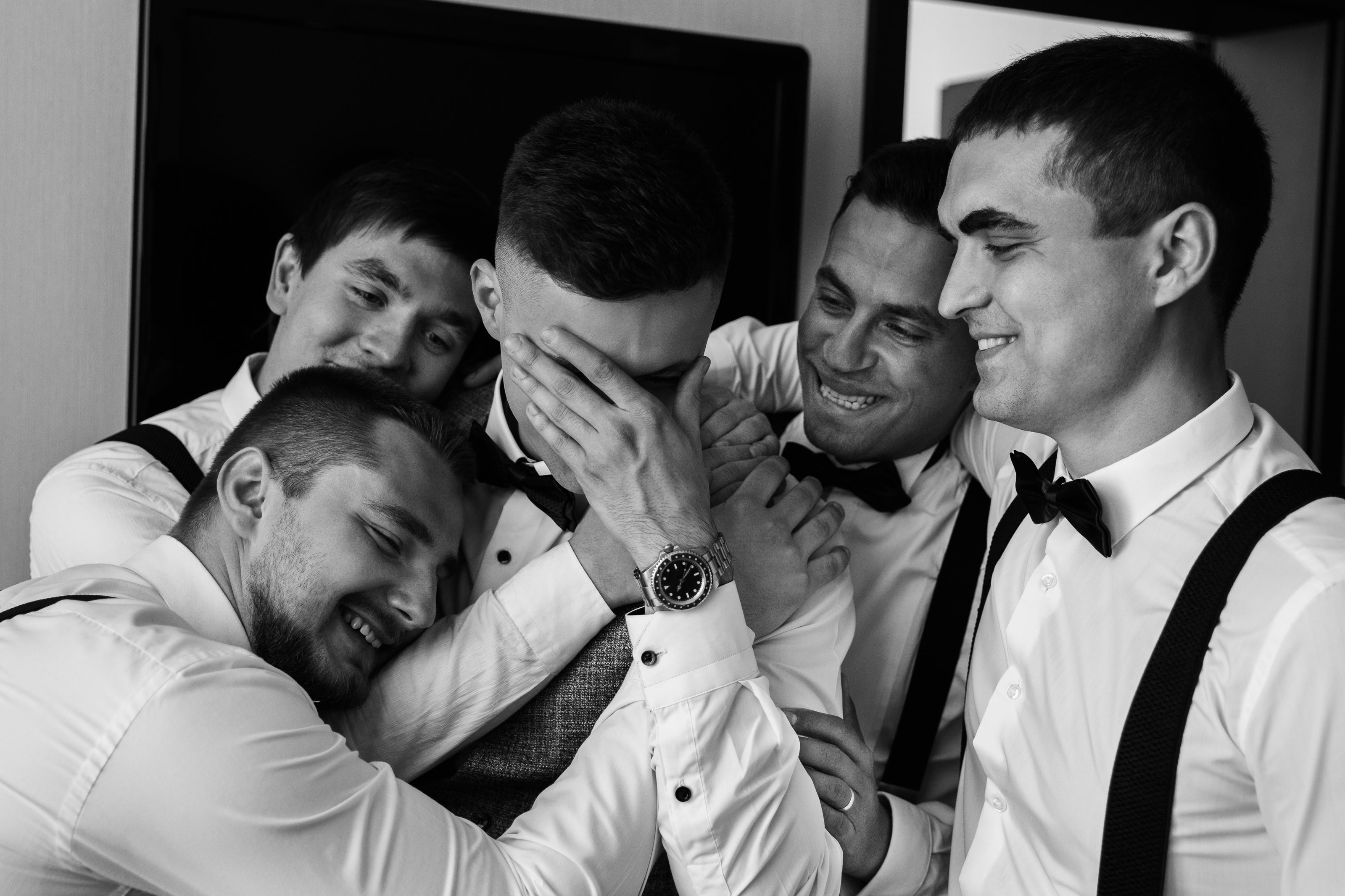 Groomsmen wearing black bow ties and suspenders in a group hug with groom in the middle hiding his face with his watch hand